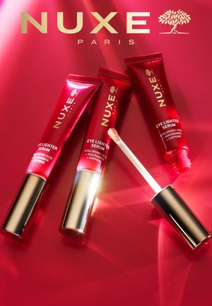 Tre tubetti rossi di siero illuminante per occhi Nuxe con tappo dorato su una superficie rossa, uno aperto con applicatore che mostra un siero beige chiaro.