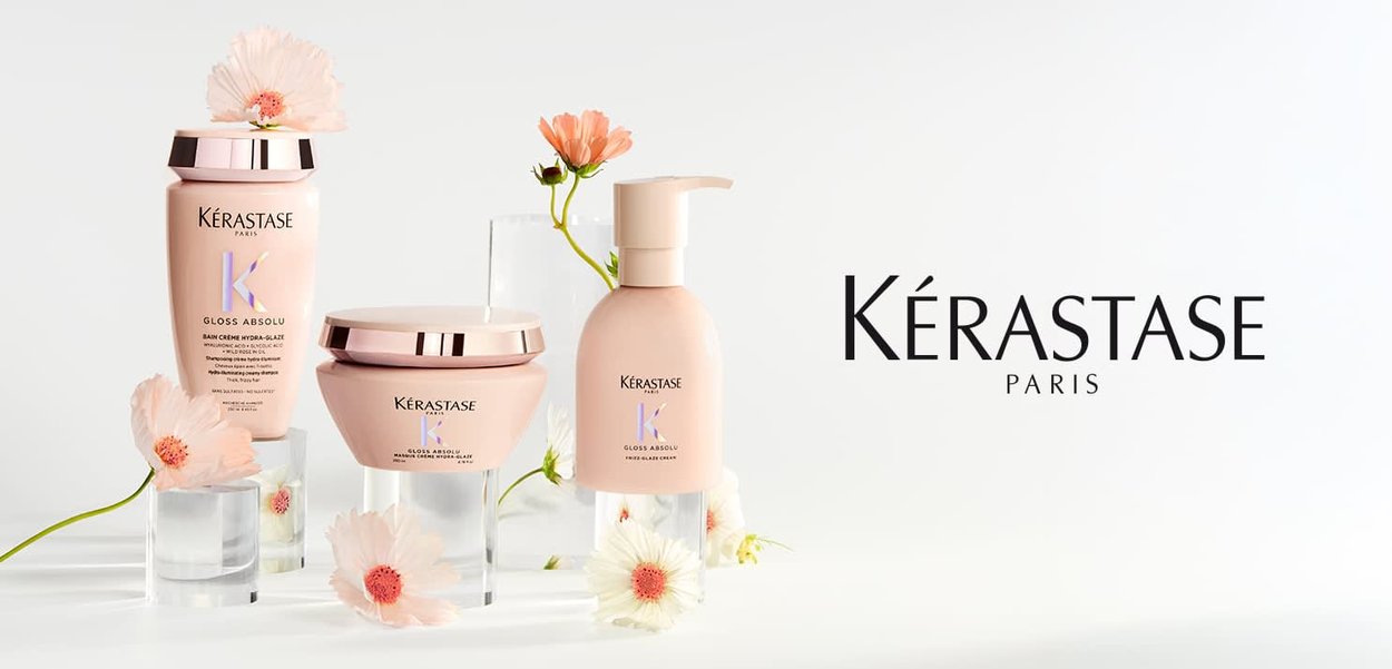 Set de cuidado capilar Kérastase Gloss Absolu con champú, crema y sérum rodeado de flores de durazno sobre fondo blanco.