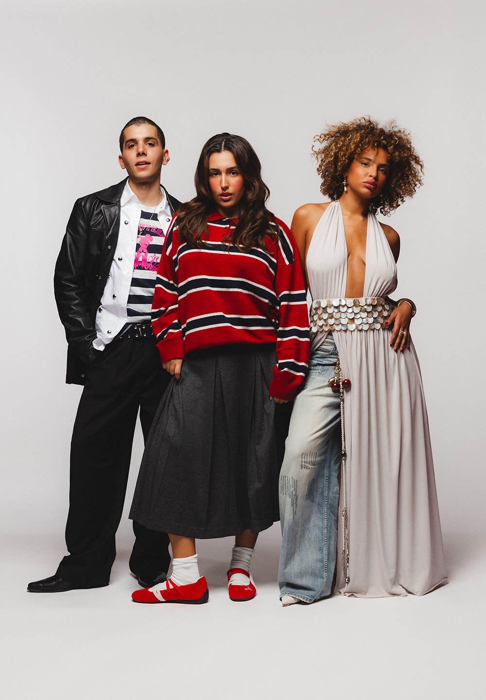 Trois personnes se tenant devant un fond clair. La première porte une veste en cuir noire et un t-shirt graphique. La deuxième porte un pull surdimensionné à rayures rouges et une jupe grise. La troisième arbore une robe blanche sans manches ornée de sequins et d'accents en denim.