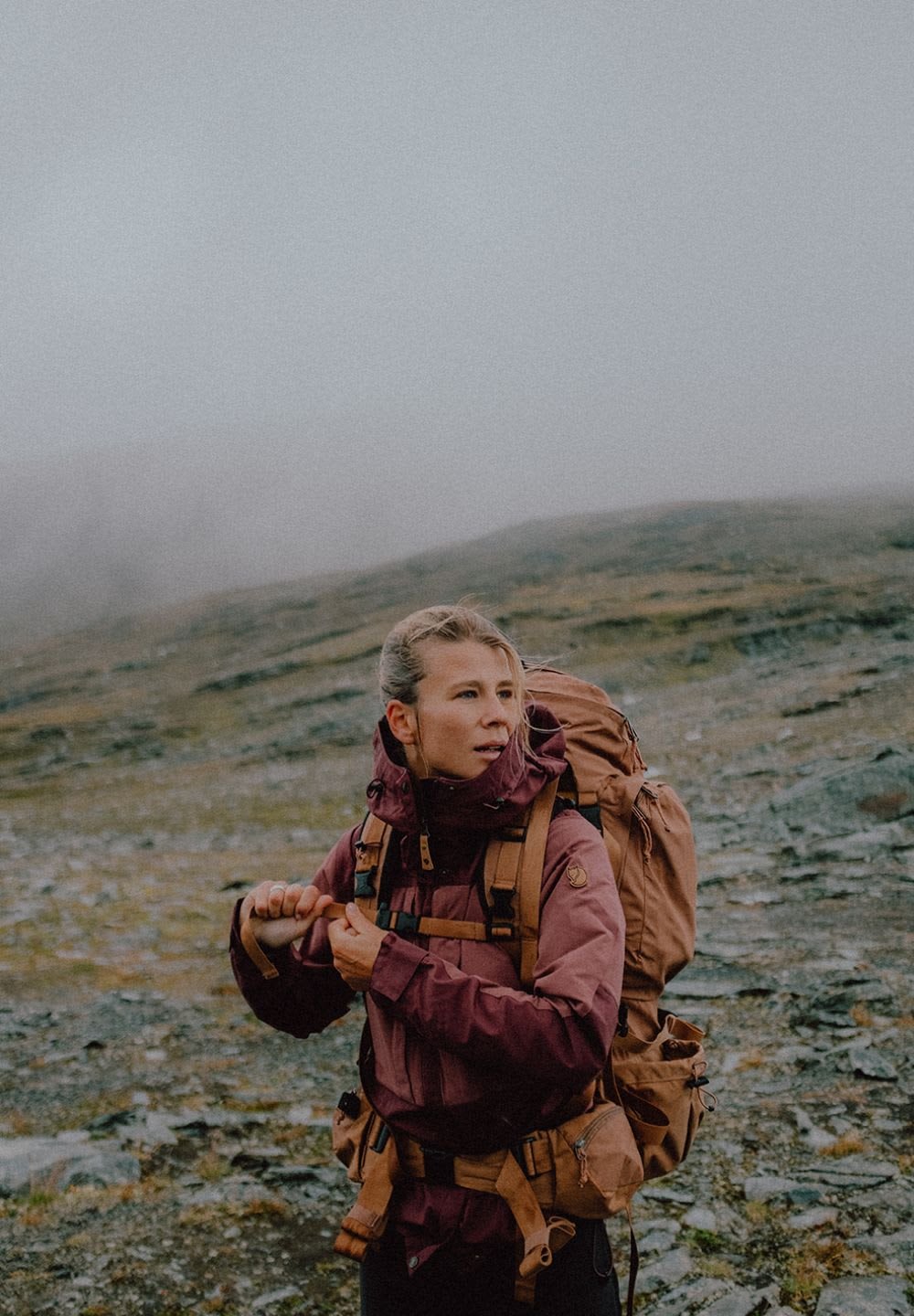 Bordeauxfärgad vandringsjacka med hög krage, brun ryggsäck med flera fickor, blöt stenig terräng och molnig himmel i bakgrunden.