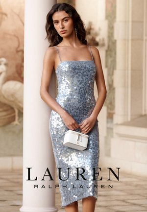 Mujer con un vestido tipo slip plateado de lentejuelas que sostiene un pequeño bolso blanco, de pie junto a una columna blanca en un entorno arquitectónico clásico.
