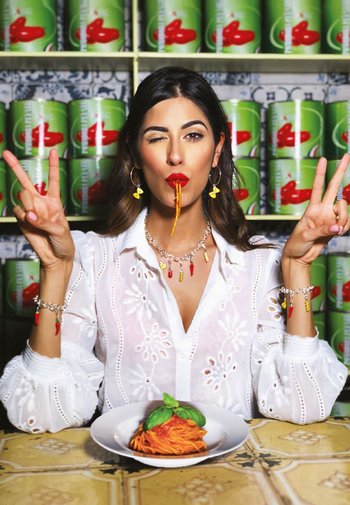 Une femme portant une blouse blanche brodée tient une assiette de spaghetti garnie de basilic. En arrière-plan, on aperçoit des tomates en conserve vertes. Elle a de longs cheveux bruns et des accessoires, dont des boucles d'oreilles en forme de piments rouges.