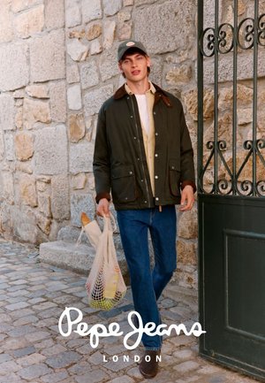 Giovane uomo con giacca casual e jeans che cammina su una strada di pietra portando una borsa di rete con la spesa vicino a un cancello in ferro battuto.