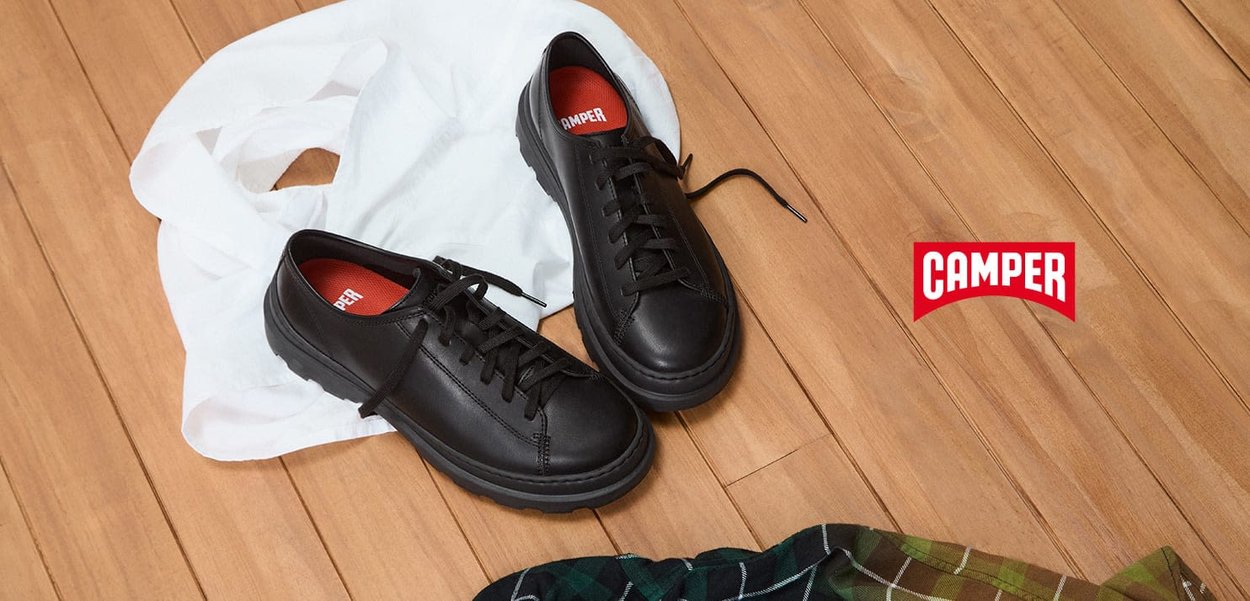 Schwarze Leder-Sneaker mit runder Zehenpartie, flachen Schnürsenkeln und Gummisohlen. Rote Innensohle mit "CAMPER"-Logo. Auf einer Holzoberfläche präsentiert.