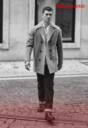 Grauer doppelt geknöpfter Mantel über einem weißen Hemd, kombiniert mit dunklen Jeans und schwarzen Schnürstiefeln, vor einem Kopfsteinpflasterstraßen-Hintergrund.