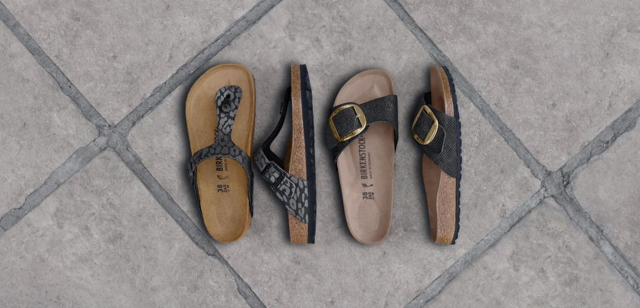 Due paia di sandali Birkenstock con motivi neri e suole in sughero posati su pavimento piastrellato grigio.