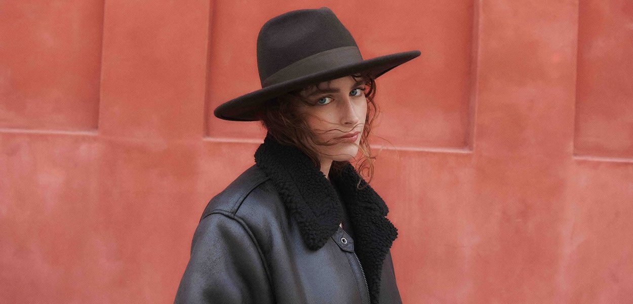 Black leather jacket with a shearling collar, worn over a black top. The person also wears a wide-brimmed brown hat against a textured pink wall.