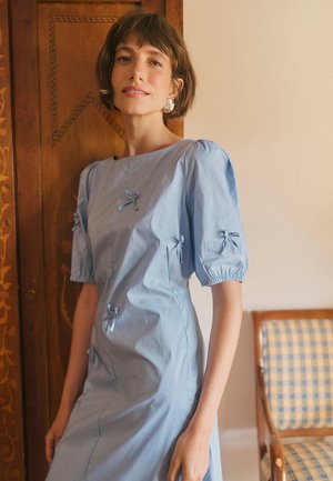 Frau mit kurzen braunen Haaren, die ein hellblaues Kleid mit kleinen Schleifen trägt, steht drinnen in der Nähe einer Holztür und eines karierten Stuhls.