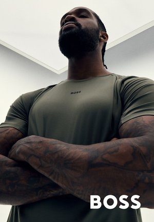 Hombre con tatuajes cruza los brazos, lleva una camiseta BOSS de color verde oliva, visto desde un ángulo bajo contra un fondo de techo brillante.