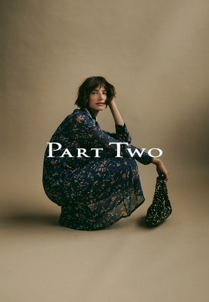 Frau mit kurzem, dunklem Haar hockt in einem langen, marineblauen Blumenkleid und hält eine schwarze, mit Pailletten besetzte Tasche, Text "Teil Zwei" in der Mitte.