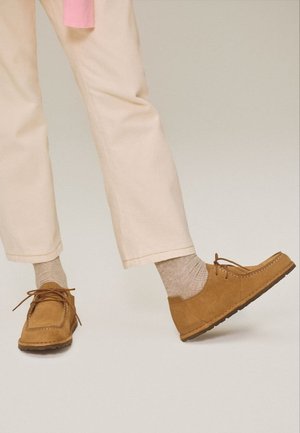 Person trägt hellbeige Hosen, beige Wildleder-Schnürschuhe und beige Socken und steht mit einem leicht angehobenen Fuß vor einem neutralen Hintergrund.