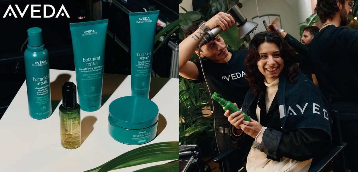 Aveda branded hair salon scene with a woman in a black jacket, holding a green spray bottle, smiling while styled by a professional.