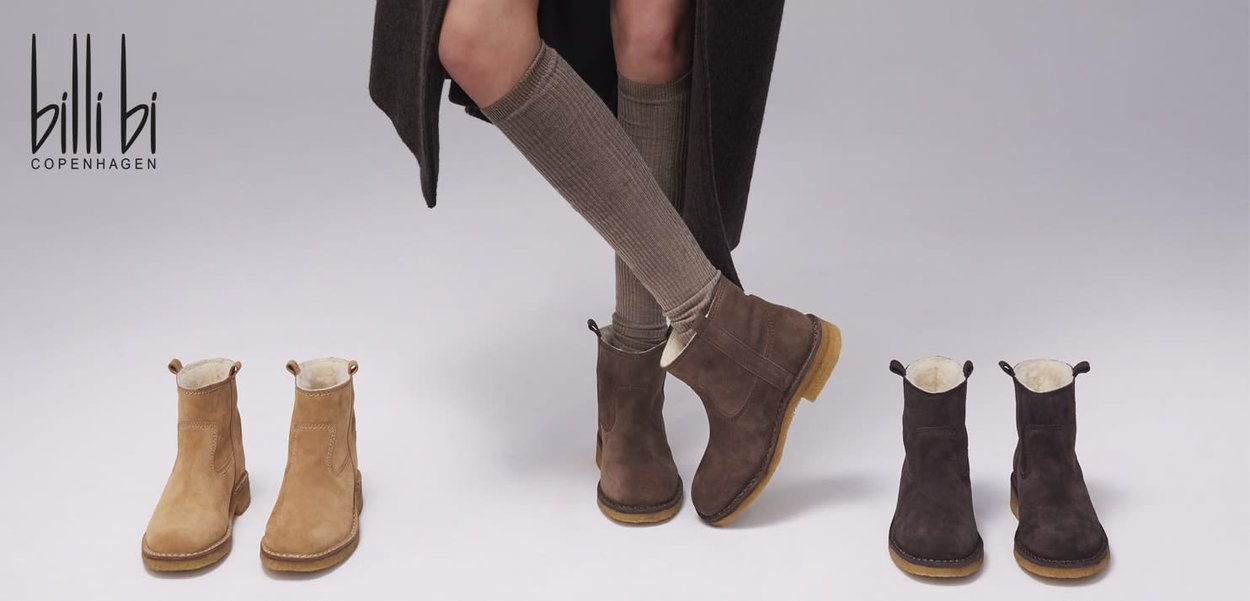 Stivali ankle in suede color tan e marrone scuro con morbido fodero in pelliccia, linguette di presa e suole testurizzate. Due paia esposti accanto alle gambe di un modello.
