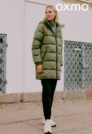 Olivengrøn puffjakke med hætte, lynlåslommer og elastiske detaljer. Kombineret med sorte bukser og hvide sneakers. Urban baggrund.