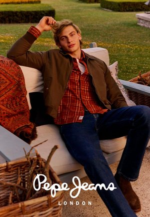 Giovane uomo in camicia a quadri, giacca marrone e jeans che si rilassa su un divano da esterno con cuscini e coperta in un giardino.