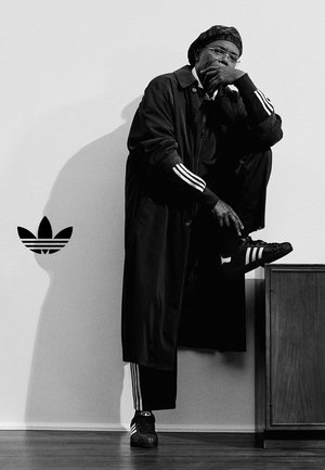 Mężczyzna ubrany w dres Adidas, beret i okulary pozujący z jedną nogą uniesioną na szafce na tle jasnej, jednolitej ściany z logo Adidas.