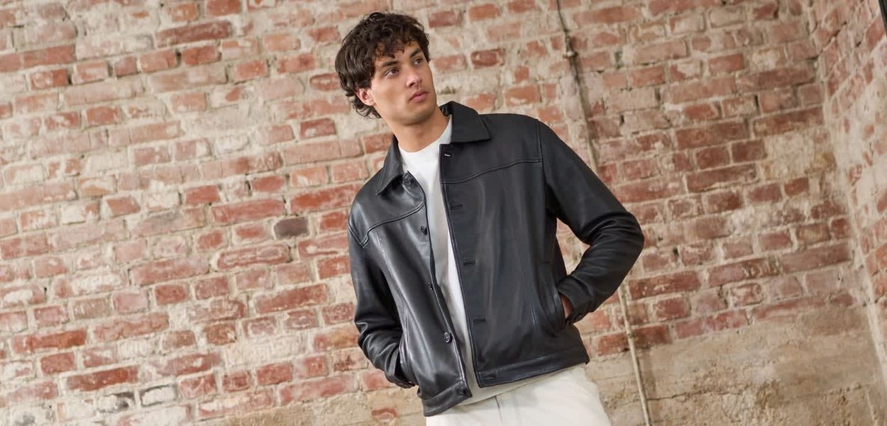 Giovane uomo con giacca di pelle nera sopra una camicia bianca, in piedi con le mani nelle tasche davanti a un muro di mattoni a vista.