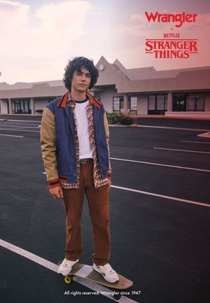 Veste bicolore bleu et beige, chemise à carreaux, t-shirt blanc, pantalon marron et baskets blanches. Debout sur un skateboard dans un parking.