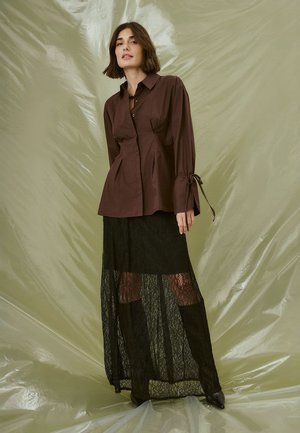 Brown collared shirt with pleats and tie accents, paired with a sheer black lace maxi skirt featuring a flared design and fabric detailing.