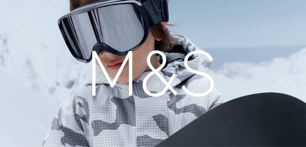 Personne portant des lunettes de ski réfléchissantes et une veste camouflage grise tient un snowboard devant un paysage montagneux enneigé.