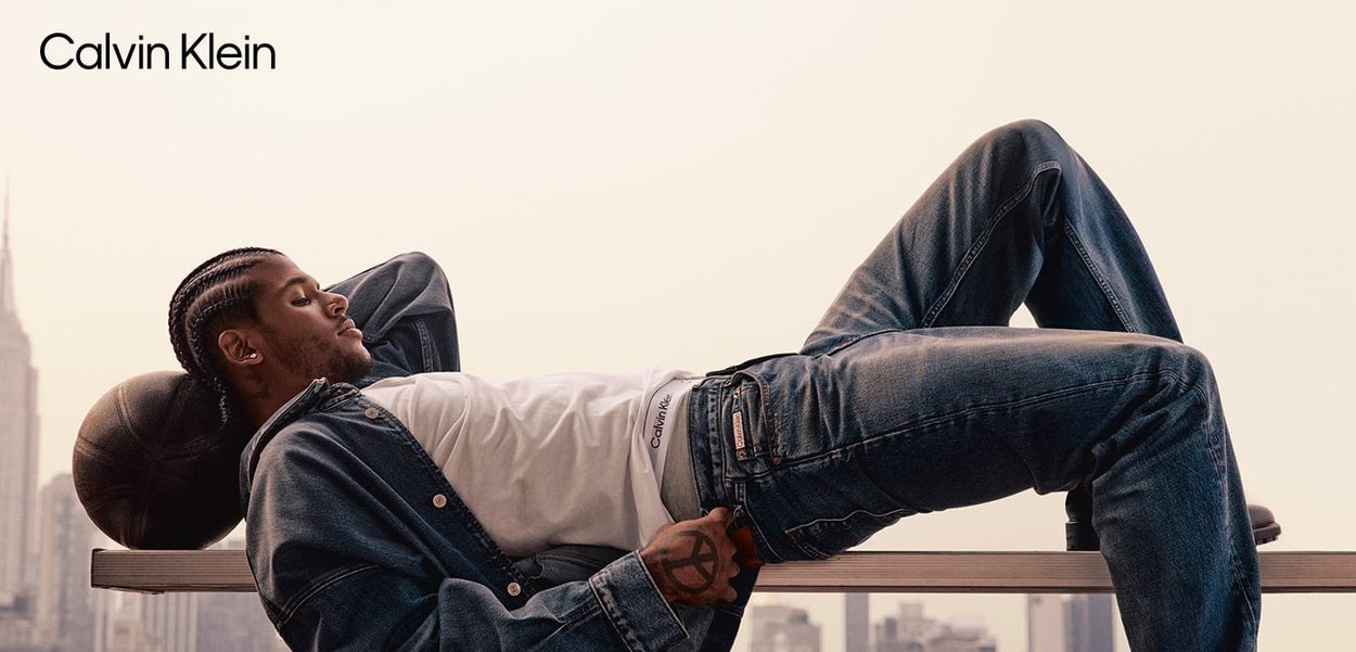 Calvin Klein Jeans aus blauem Denim, getragen mit einem weißen T-Shirt. Die Person lehnt sich auf einer Kante zurück, mit einem Basketball in der Nähe; Stadtansicht im Hintergrund.