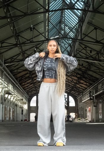 Camo-print pufferjack met een cropped zwarte graphic tee, grijze sweatpants en gele instappers; heeft lang, gevlochten haar.