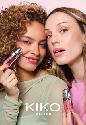 To kvinner holder leppeprodukter i slanke beholdere med sølvlokk. Den ene flasken er mørkerød, den andre myk rosa. Begge har lett sminke og pastellfargede topper.