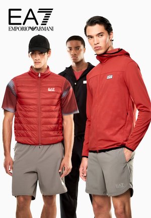 Drei Männer in EA7 Emporio Armani Sportbekleidung: zwei in roten Jacken und grauen Shorts, einer in schwarzer Jacke und rotem Shirt, posieren vor weißem Hintergrund.