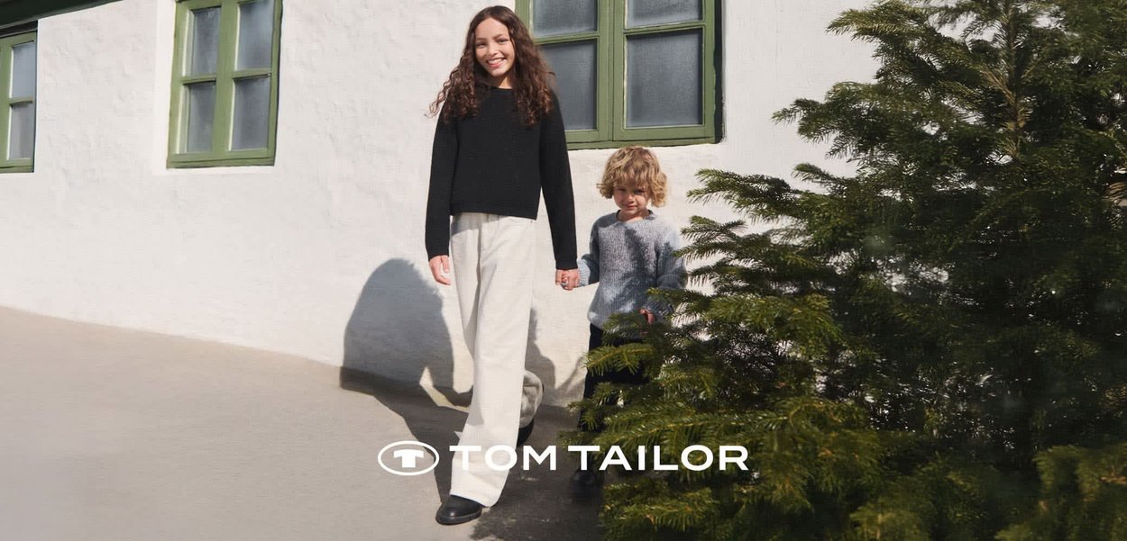 Lächelnde Frau und Kind halten im Freien bei einem grünen Fenster und einer Kiefer Händchen, beide tragen Strickpullover und Hosen.