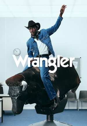 En person iført en jeansjakke og cowboyhatt rir på en mekanisk okse, plassert i et kontormiljø med blå teppe og stoler.