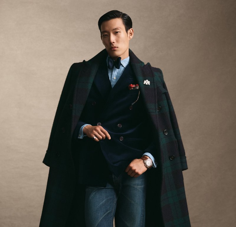 Manteau à carreaux noir et vert superposé sur un blazer croisé bleu marine, chemise bleu clair, jeans en denim, et une montre argentée. Détail de épingle rouge et blanche.