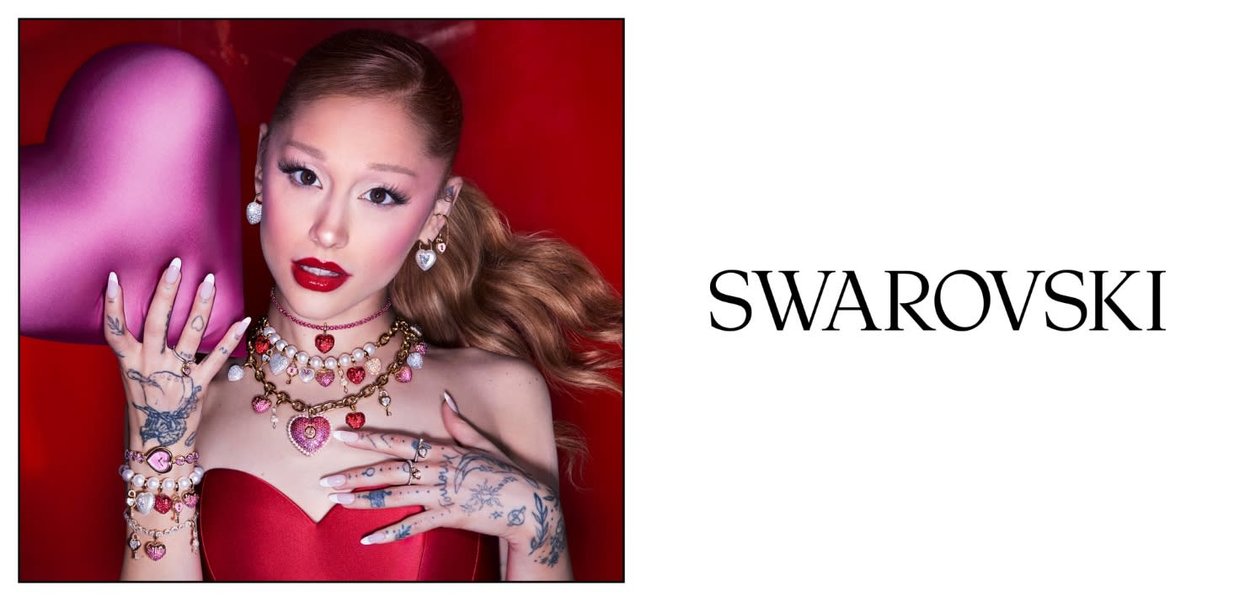 Kobieta z biżuterią w kształcie serca, tatuażami i czerwoną sukienką trzyma różowe serce na czerwonym tle obok logo Swarovski.