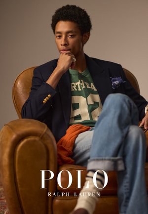 Blazer marine avec des boutons dorés porté sur un sweatshirt graphique vert et un jean en denim. Assis dans une chaise en cuir marron avec un objet orange drapé.