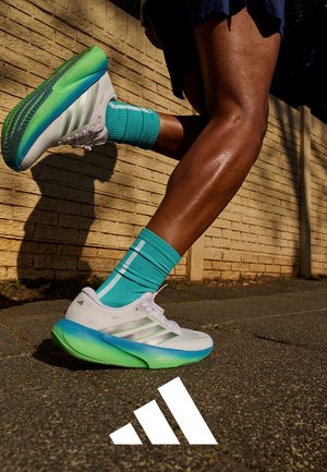 Løperens ben i turkise sokker og hvite-grønne Adidas-sko på en asfaltsti ved siden av en murvegg, med Adidas-logo på bakken.
