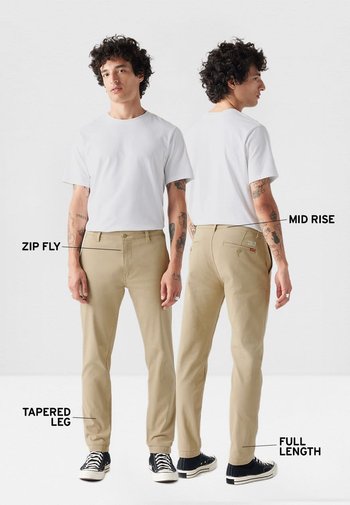 Pantalones beige de corte entallado, con cintura de talle medio y cremallera, diseñados con un corte de longitud completa. Presentados en un modelo que lleva una camisa blanca.