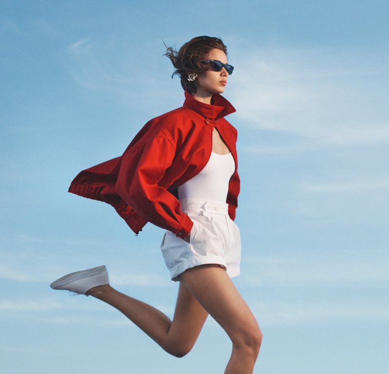 Joven mujer con pantalones cortos blancos, camiseta de tirantes blanca, chaqueta roja y gafas de sol caminando con paso decidido contra un fondo de cielo azul despejado.