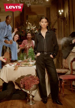 Chemise noire avec passepoil blanc, jean foncé et silhouette ajustée portée par un mannequin dans une pièce décorée avec une table à dîner et plusieurs personnages.