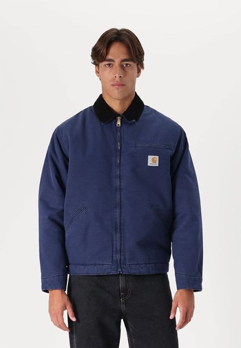 Marineblauwe jas met een zwarte kraag, ritssluiting en zijzakken. Heeft een klein merkembleem op het linkerborstgedeelte.