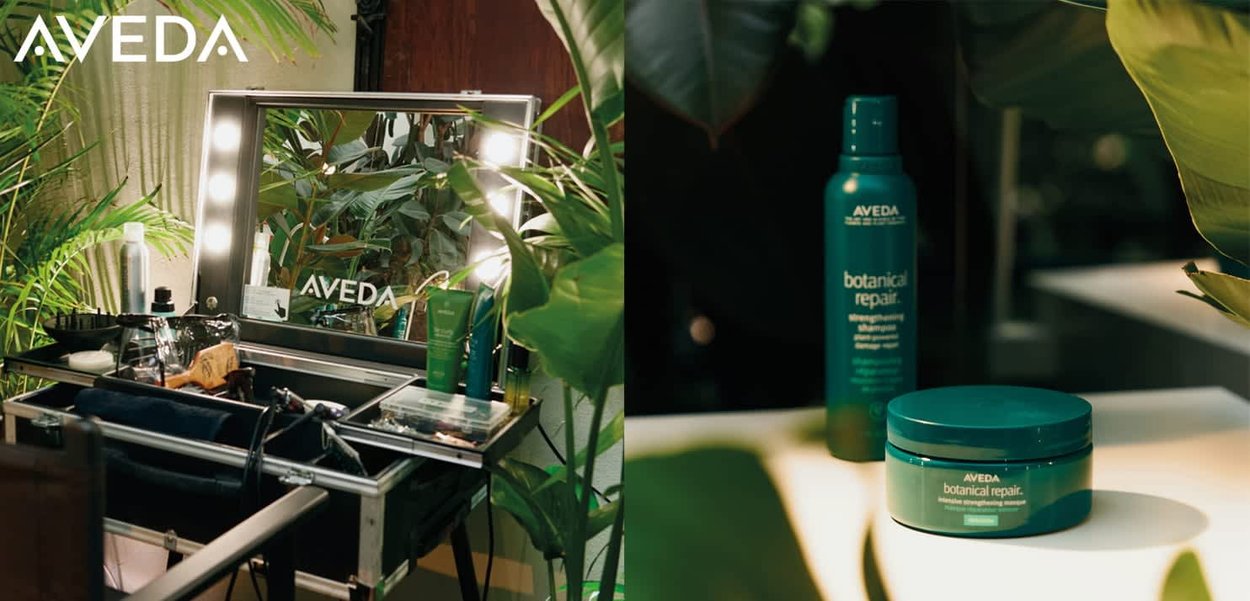 Spiegel mit Beleuchtung, grüne Haarpflegeprodukte und Werkzeuge auf einem schwarzen Wagen, umgeben von üppigen Pflanzen. Behälter in Türkis mit weißen Etiketten.