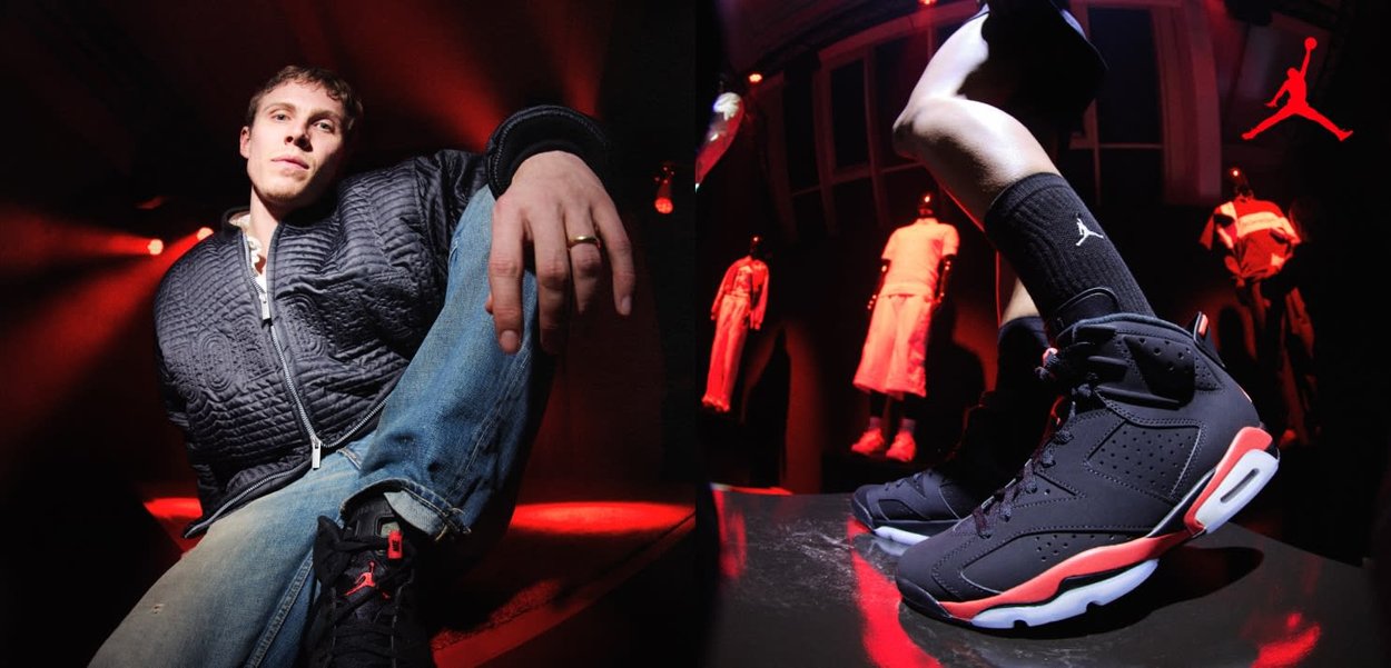 Jeune homme en vêtements décontractés assis sous des lumières rouges, présentant des baskets Air Jordan noires et rouges sur un piédestal dans une pièce sombre.