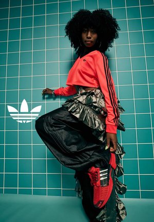 Rode cropped sweatshirt met zwarte strepen, zilveren volants broek en rode sneakers met zwarte accenten; geposeerd tegen een teal betegelde achtergrond.