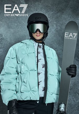Doudoune bleu sarcelle clair avec un design matelassé, portée sur un haut à motif, associée à des gants noirs, un casque et un équipement de ski.