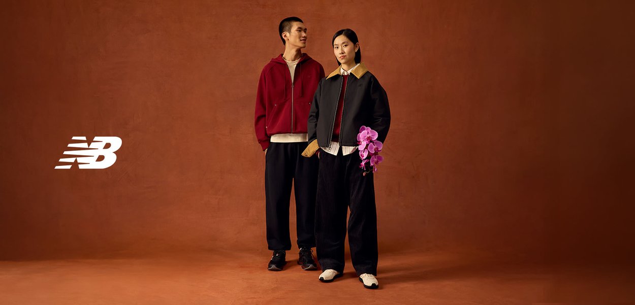 Zwei Models tragen lässige New Balance Outfits und stehen auf einem braunen Boden; eines hält eine lila Orchidee vor einem braunen Hintergrund mit NB-Logo.