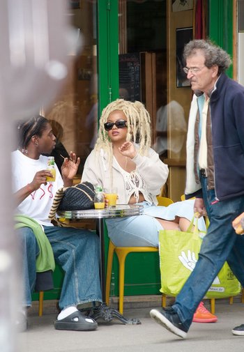 Twee mensen zitten aan een ronde tafel met drankjes; de een draagt een beige cropped hoodie en de ander een wit T-shirt, beide in casual kleding.