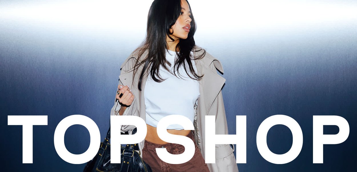 Jeune femme portant un crop top blanc, une veste beige et une jupe marron, tenant un sac à main noir, sur un fond dégradé sombre avec le texte "TOPSHOP".