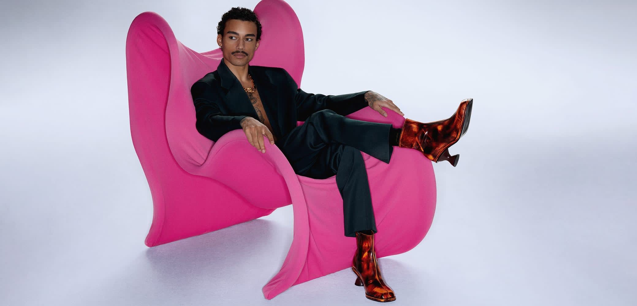Pink sculptural chair featuring a smooth, curved design. A person is seated wearing a black suit and glossy patterned brown boots, adopting a relaxed posture.