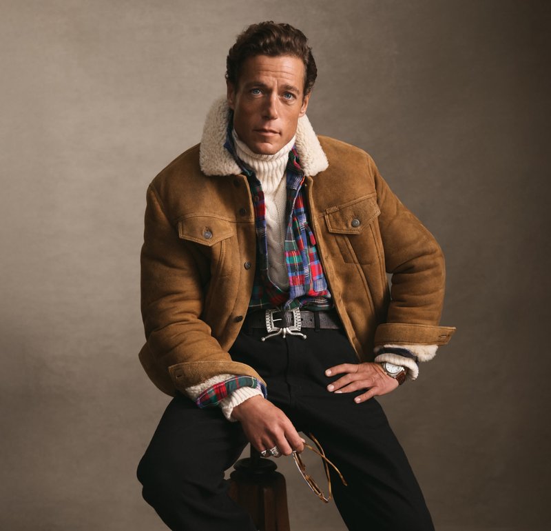 Brun mocka jacka med fårskinnskrage, lagd över en rutig skjorta och en krämfärgad polotröja. Svarta byxor och ett silverbälte med spänndetaljer.