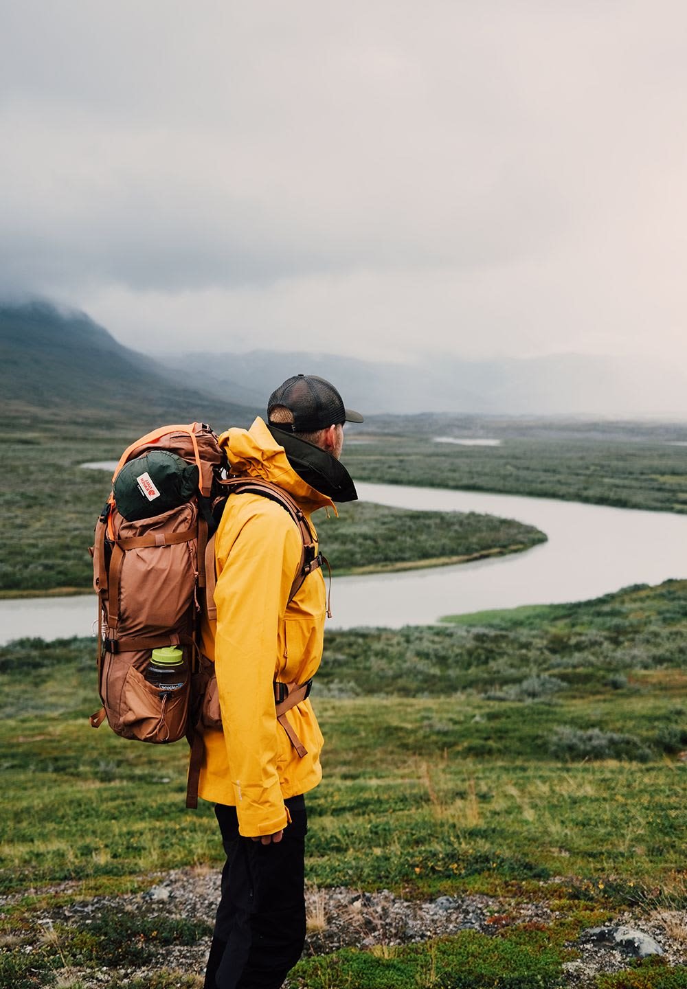 Gul vattentät jacka, beige ryggsäck med gröna accenter, svart hatt, stående i naturen med en slingrande flod och berg i bakgrunden.