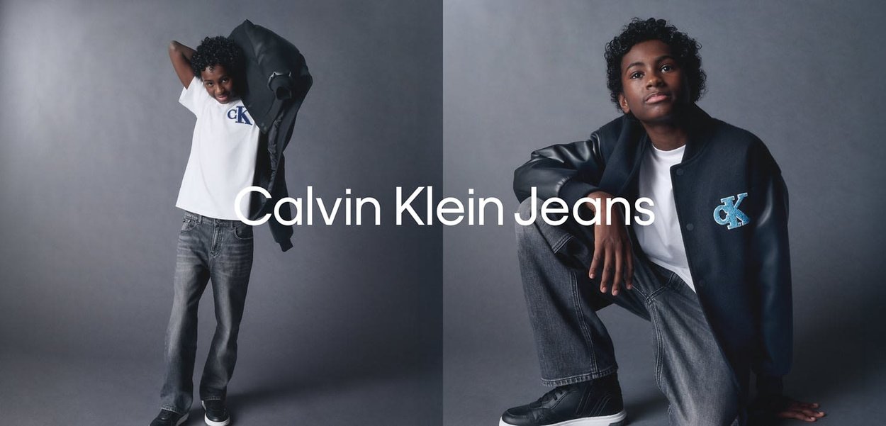 A model wears a white t-shirt with blue "CK" logo and dark denim jeans, paired with a black varsity jacket featuring blue accents.