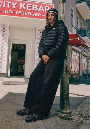 Schwarze Daunenjacke, lockere schwarze Hose und schwarze Sneaker mit weißen Akzenten. Steht an einem Straßenpoller in der Nähe eines Kebab-Ladens.